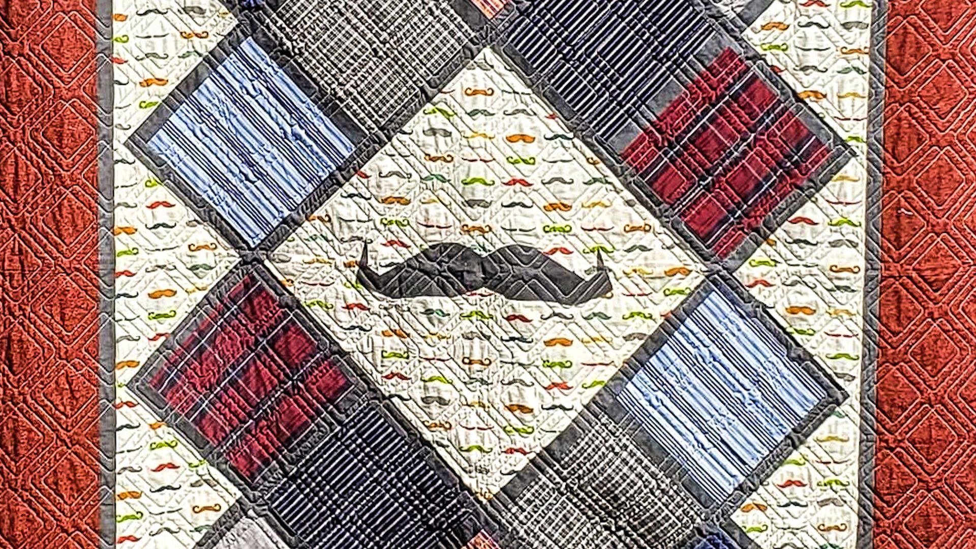 Someone holds up a quilt, with a large moustache quilted onto the middle of it