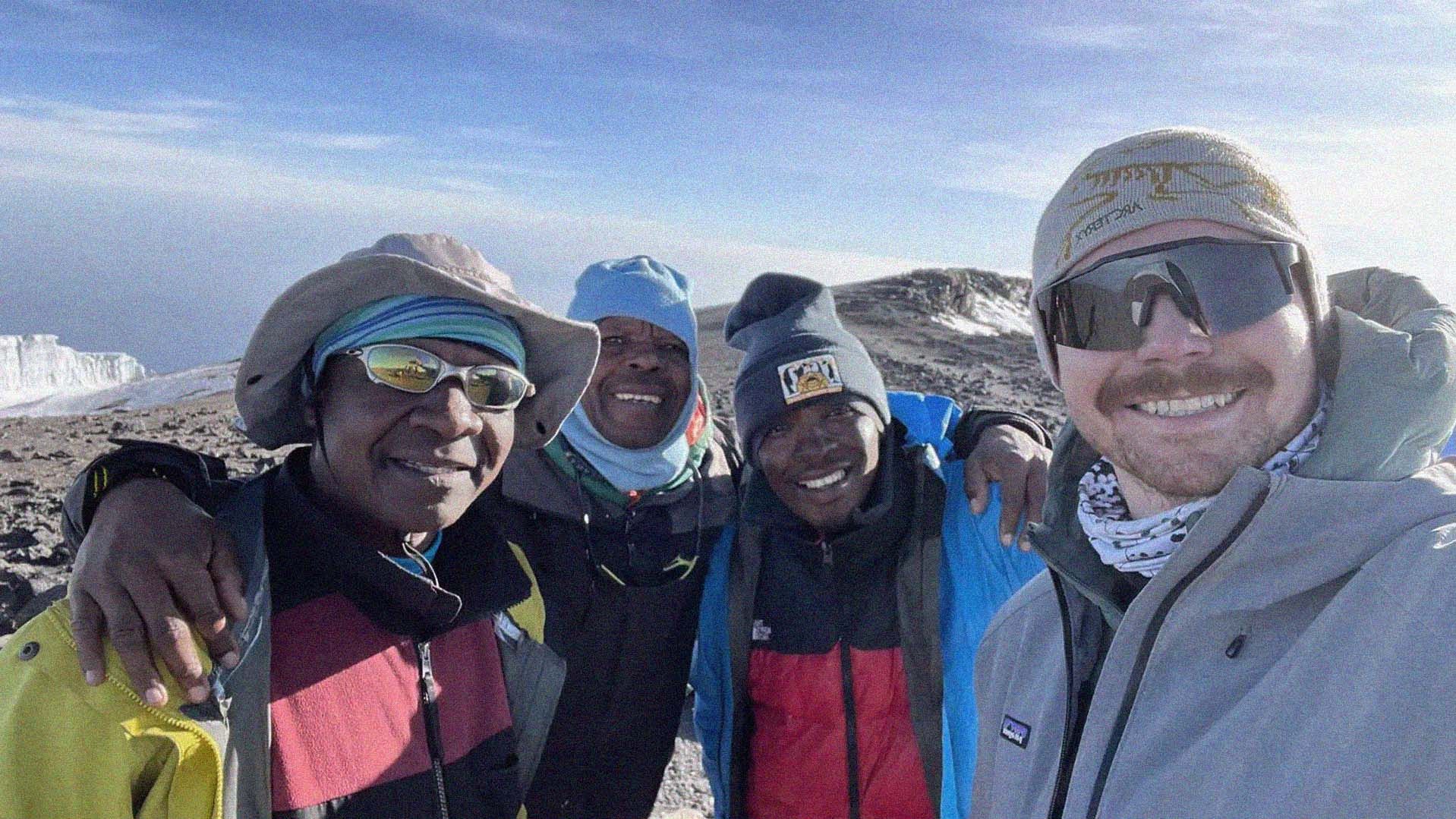 Four men in cold-weather gear, smiling to camera, while a mountain top is in the background.