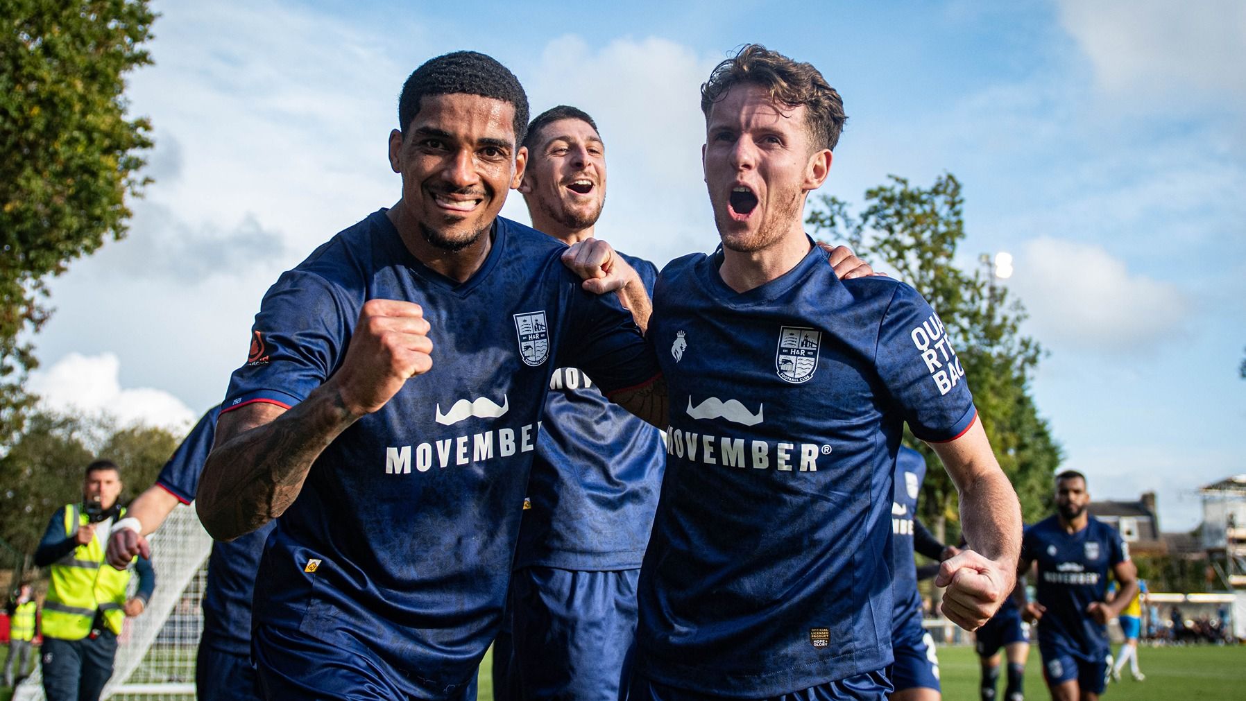 Photo of three jubilant young men on a sports pitch, wearing Movember-branded football uniforms.