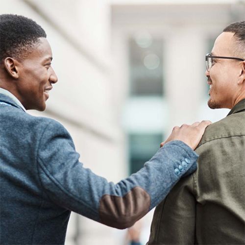 A young man placing his hand on the shoulder of another young man. The two are smiling at each other.