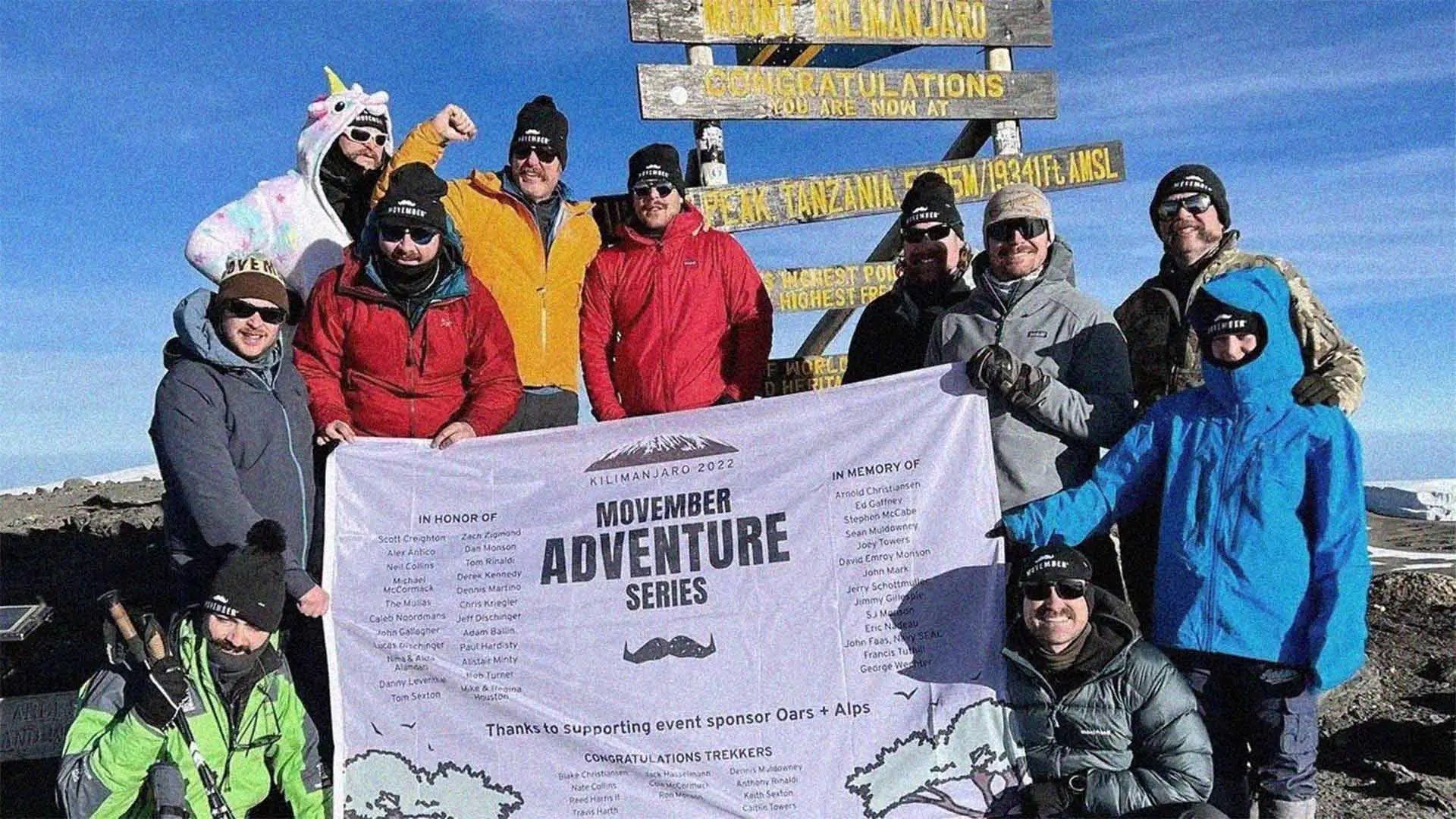 Ten Movember fundraisers in cold weather gear, atop Mount Kilimanjaro, posing with a Movember Adventure Series poster.