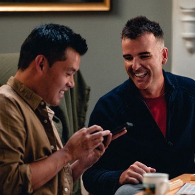 Two smiling men in casual clothes. One points out something to the other on his smartphone.