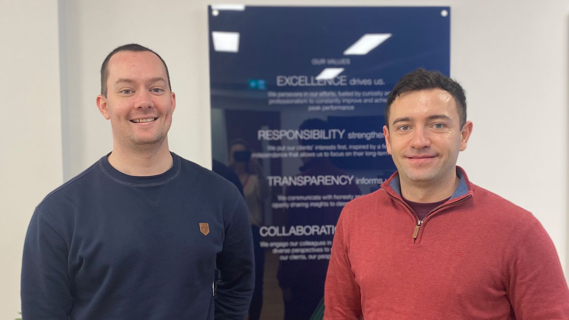 Two male colleagues post for a photo in front a poster displaying the company's values, including excellence, responsibility, transparency, and collaboration