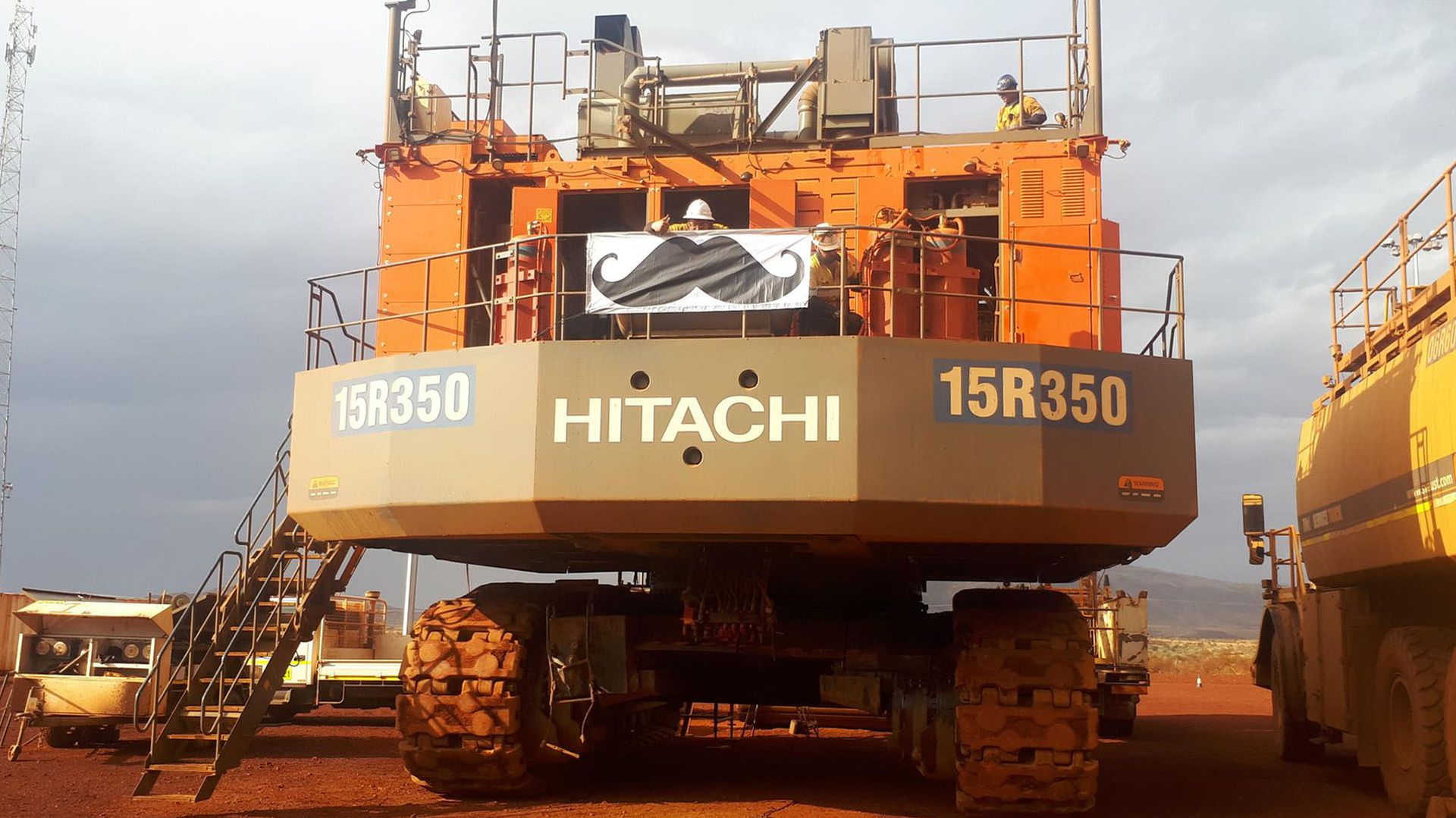 Photo of a heavy duty Hitachi earthmoving machine, adorned with a Movember moustache logo.