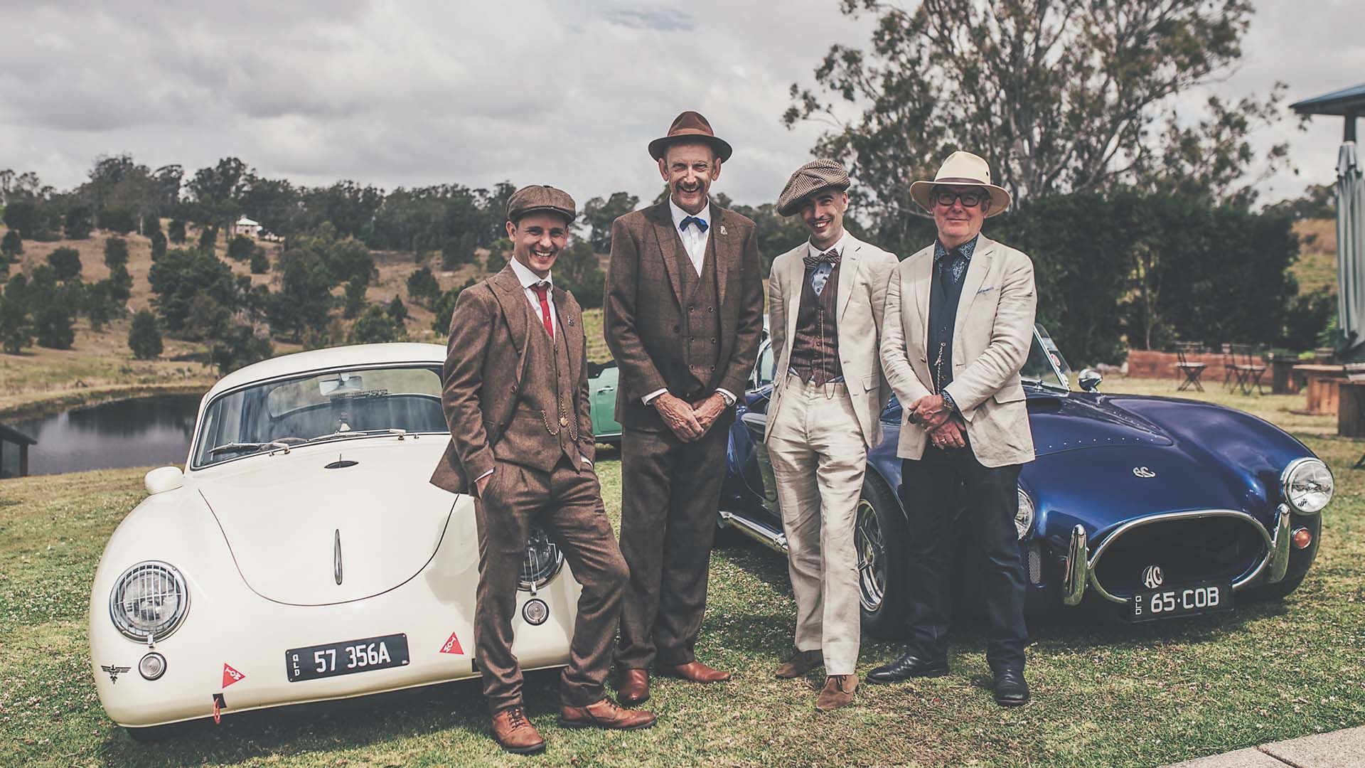 Four superbly-dressed dapper men posing in front of two classic cars.