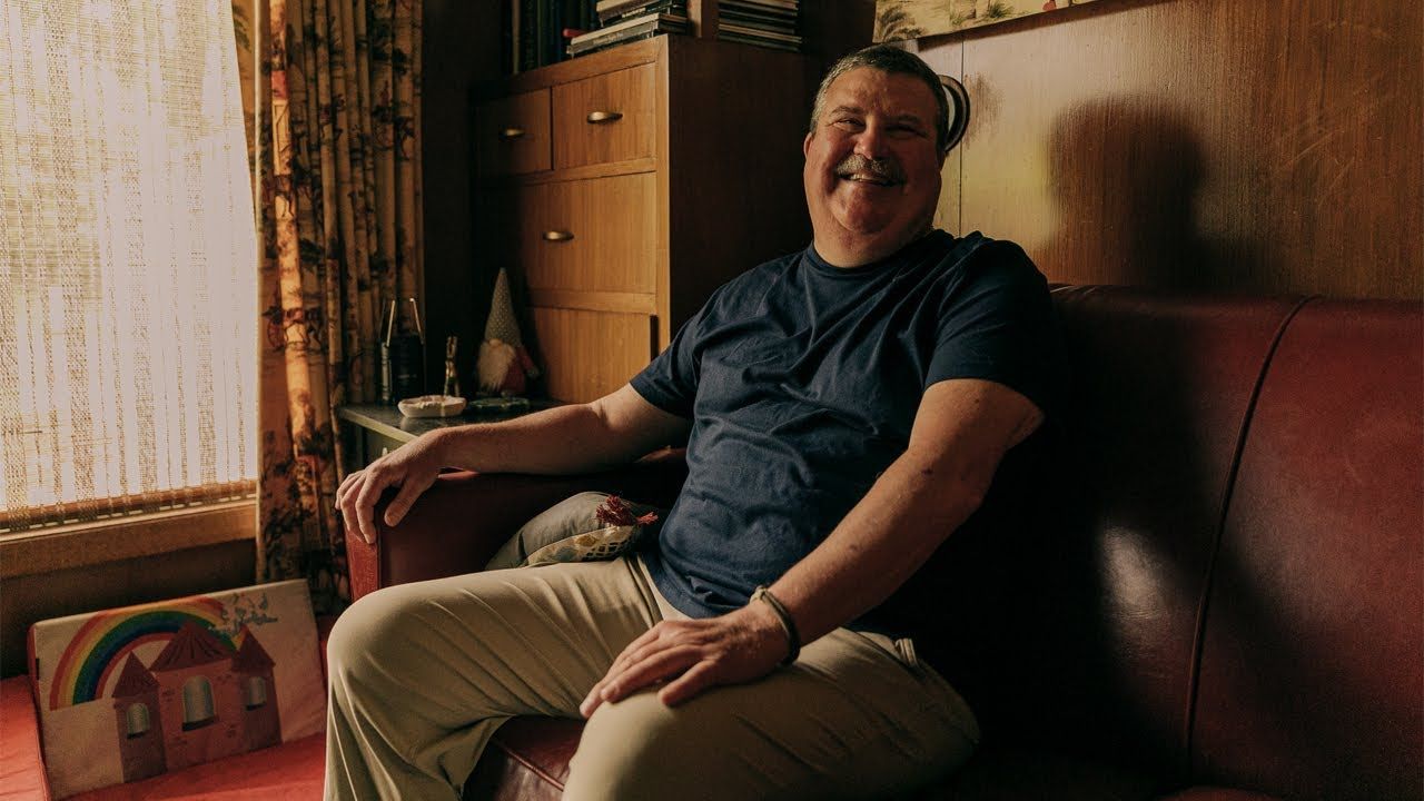 Photo of man, sitting in his living room and smiling in a jolly manner to camera.