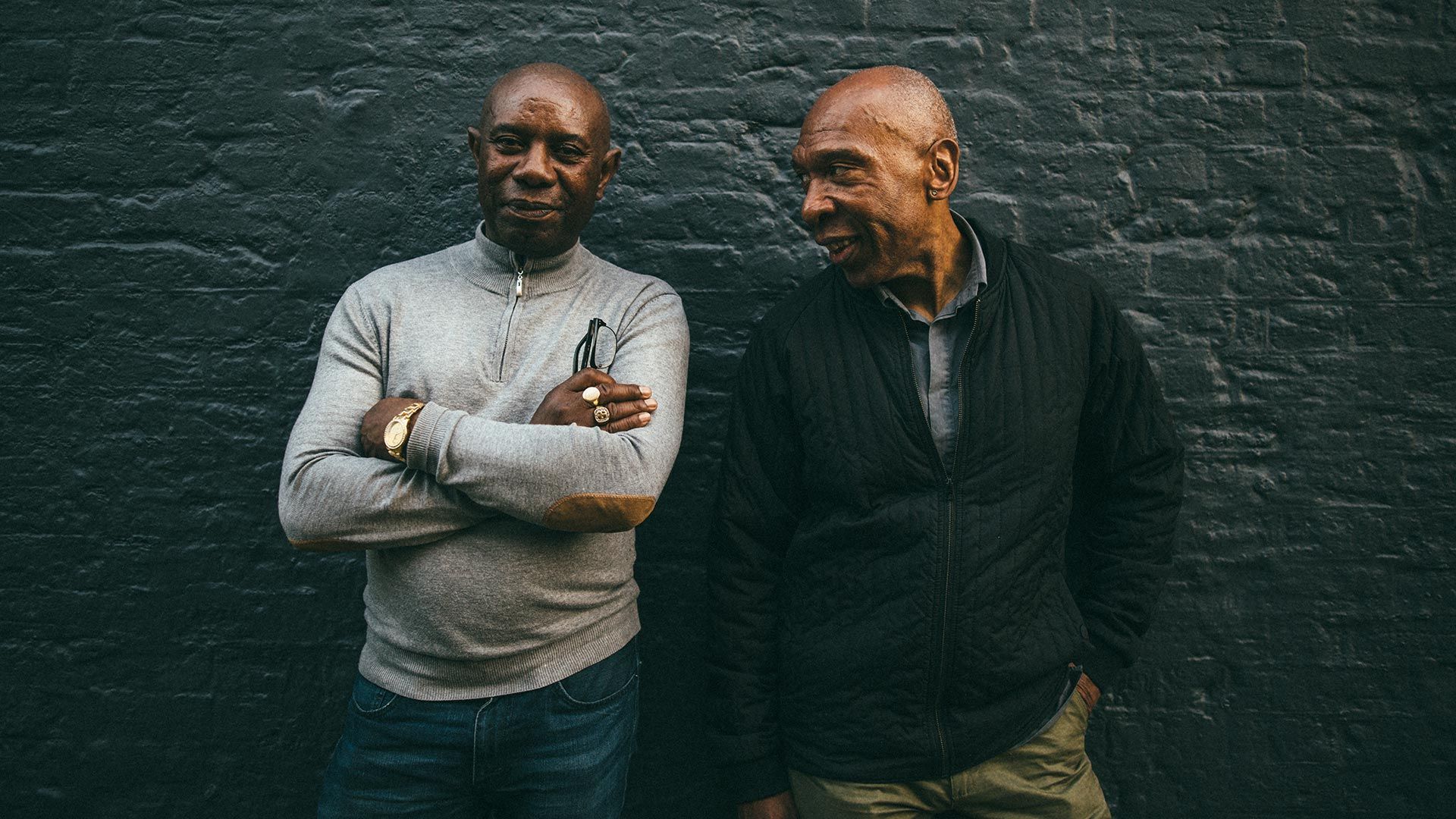 Two older male friends standing in front of a painted brick wall.