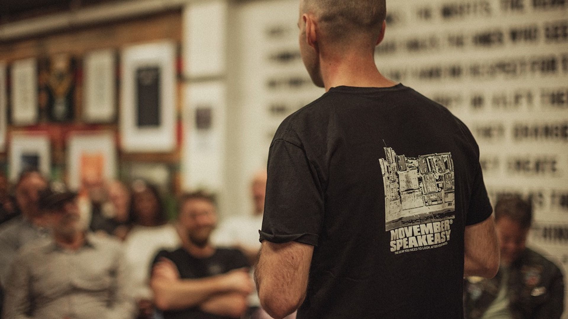 A man's back is turned to us as he presents to a full room. The back of his shirt says "Movember SpeakEasy"