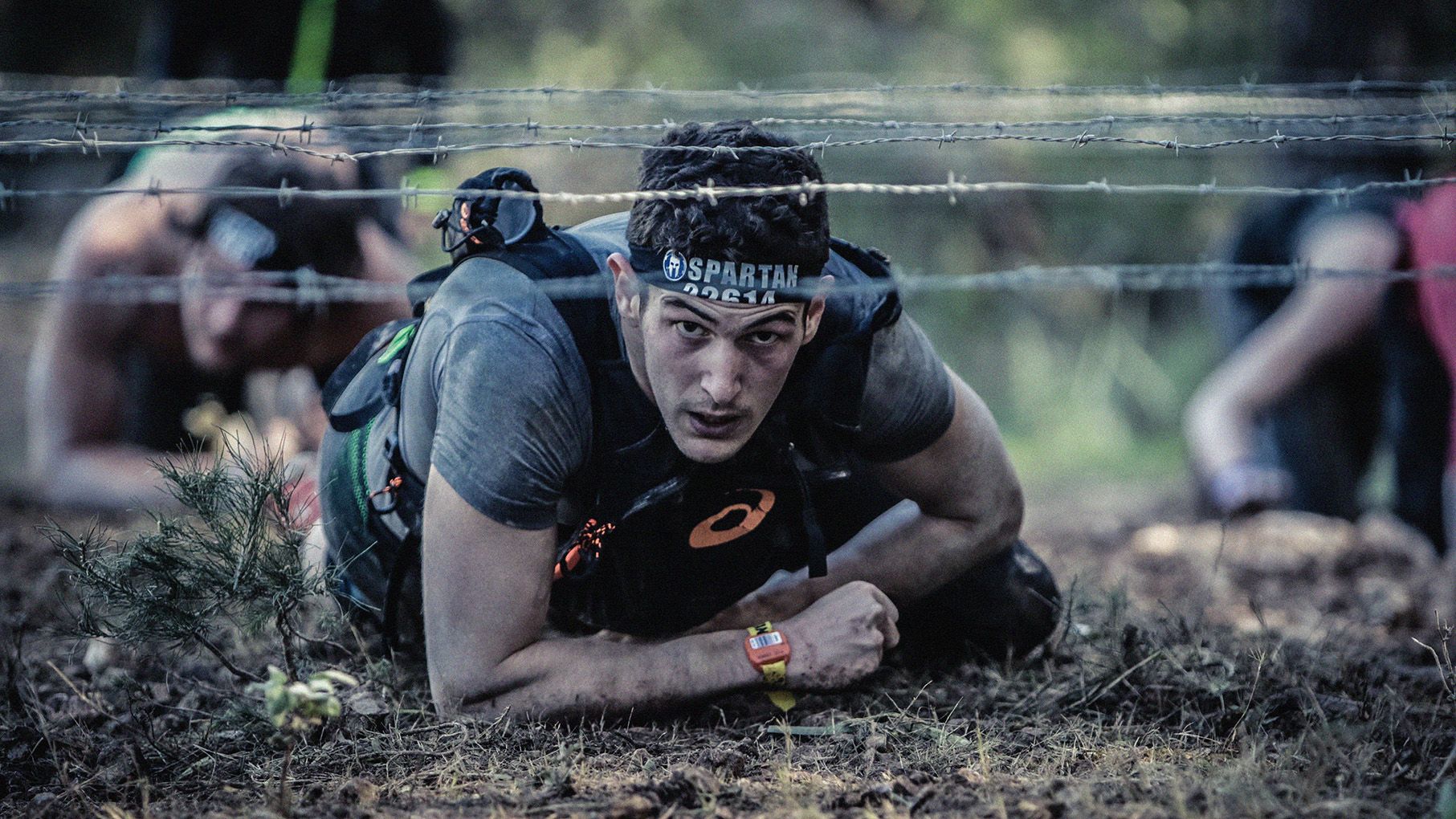 Photo of Spartan Races competitor, crawling forward on ground underneath a barbed wire obstacle.