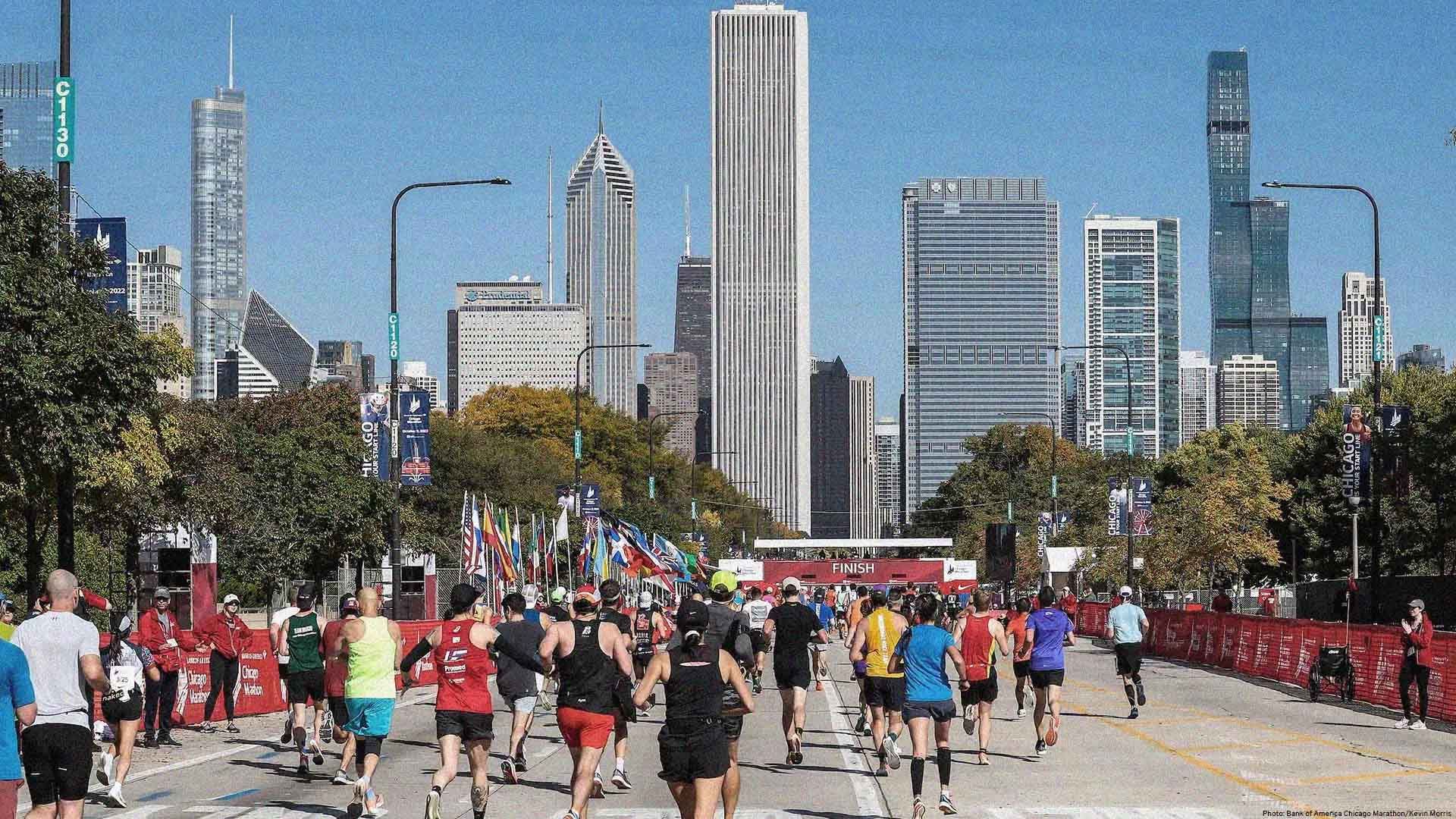 Wide photo from the rear of dozens of runners competing in the Chicago Marathon. In the background, tall skyscrapers form an impressive skyline.