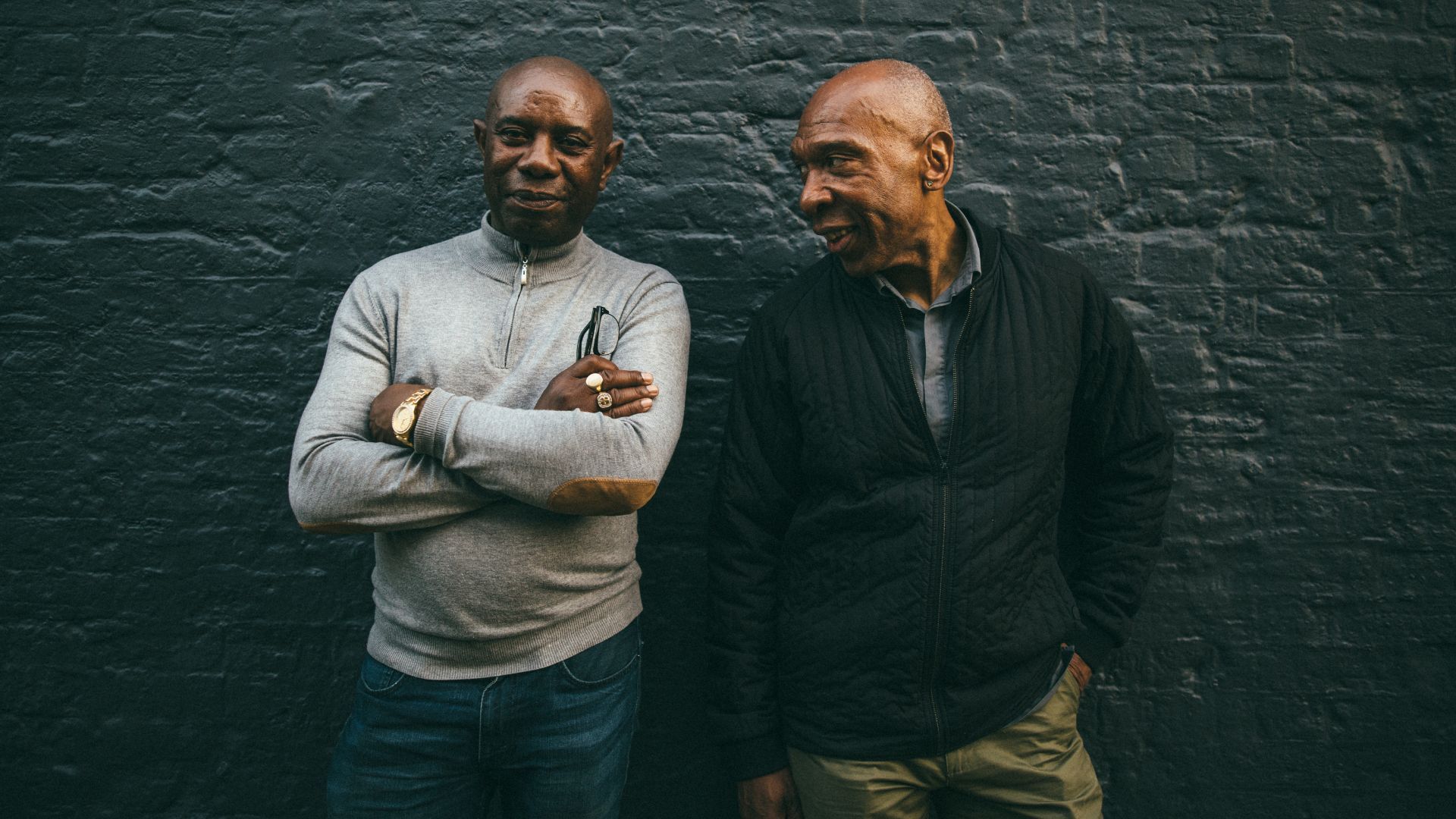 An image of two men standing together in front of a charcoal brick wall.