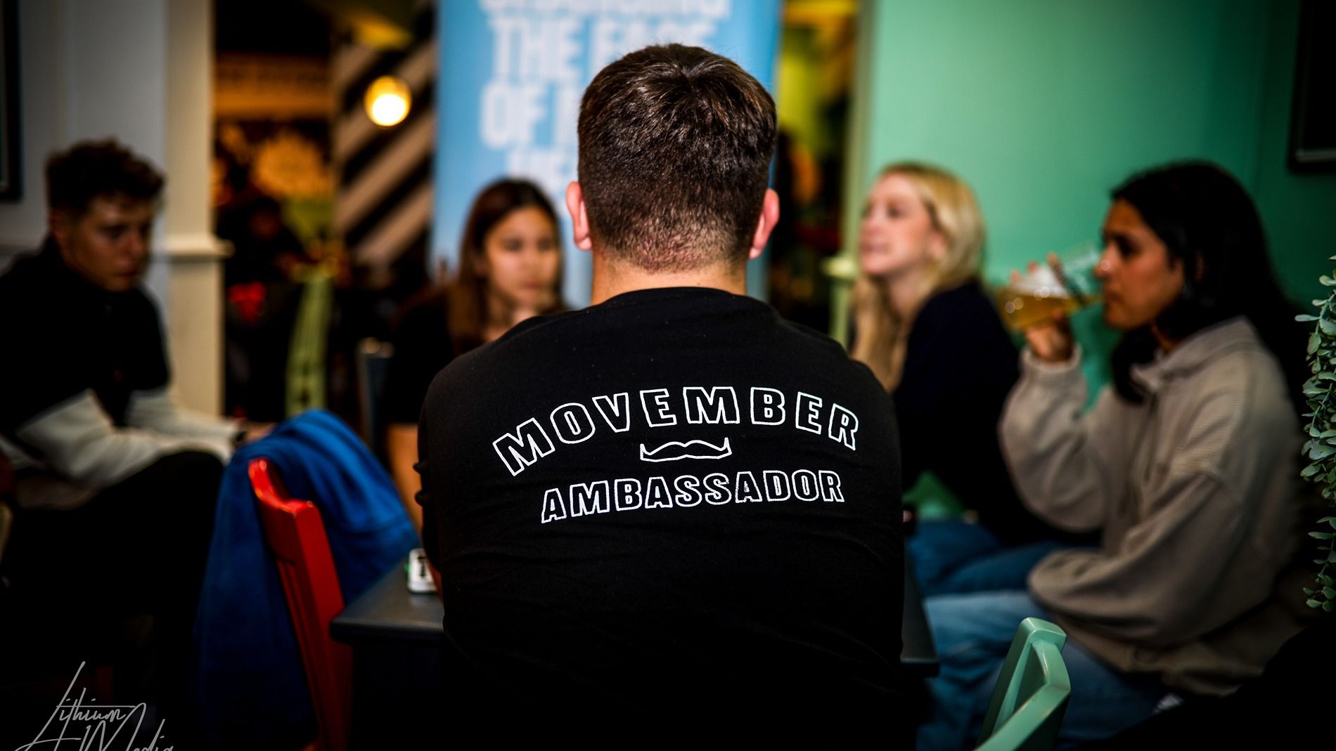 Photo of young people at a party. One wears a shirt that says "Movember Ambassador".
