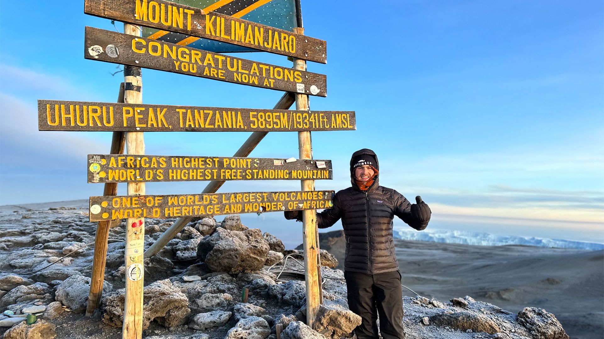 A bundled up young Mo Bro poses at the summit of Mount Kilimanjaro, with an epic vista behind him. The sign reads "Congratulations you are now at Uhuru Peak"