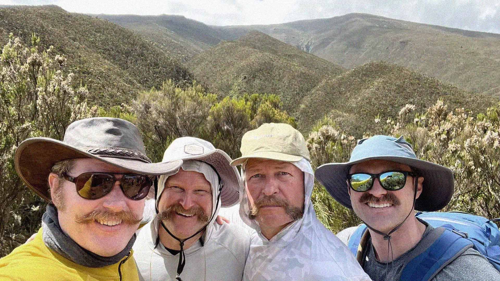 Four Movember supporters, bearing magnificent moustaches, smiling to camera. Spectacular verdant hills from Africa's Mount Kilimanjaro are visible behind them.