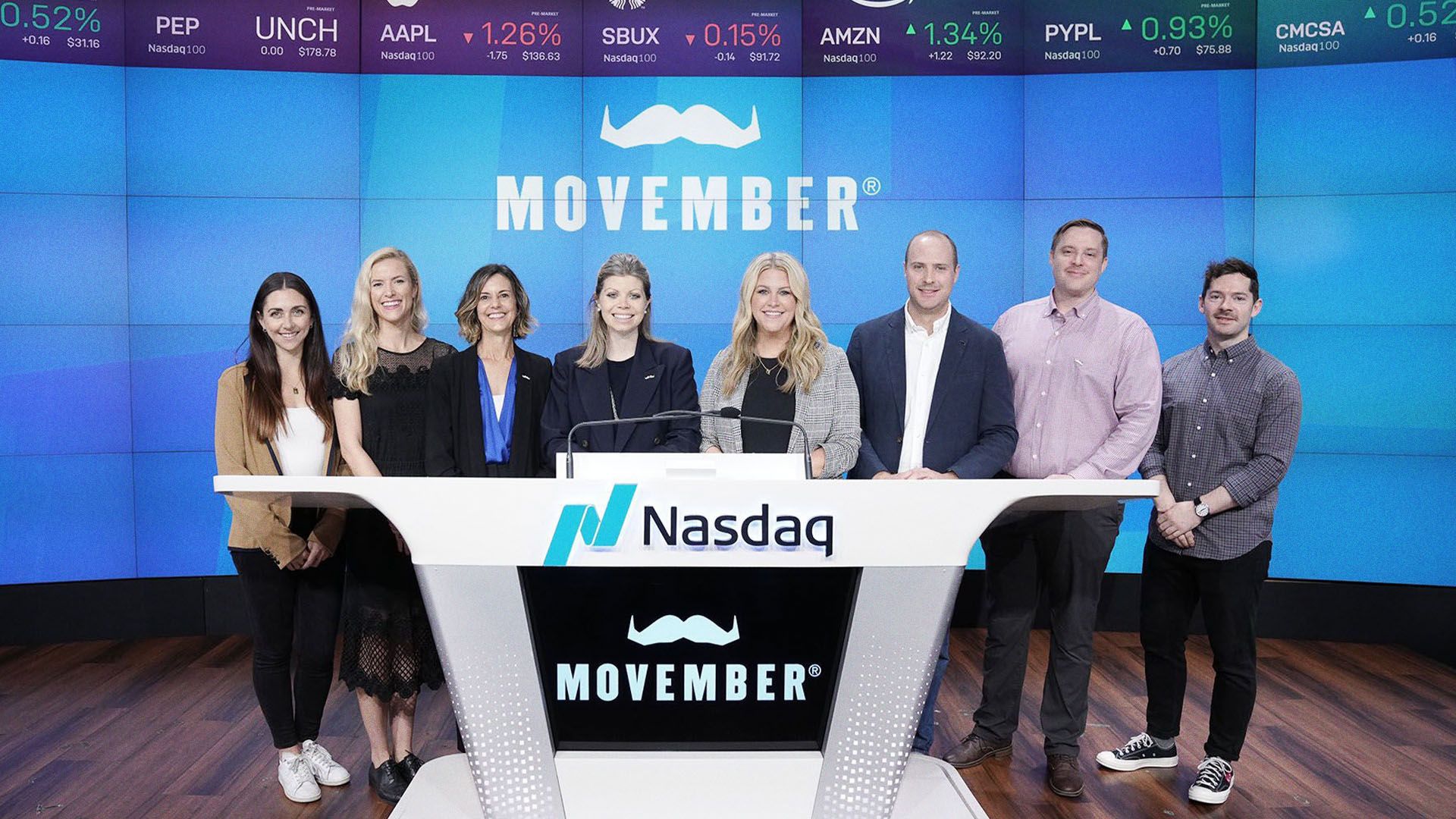 Photo of a group of smiling people from the Movember charity, excitedly about to ring the NASDAQ bell.