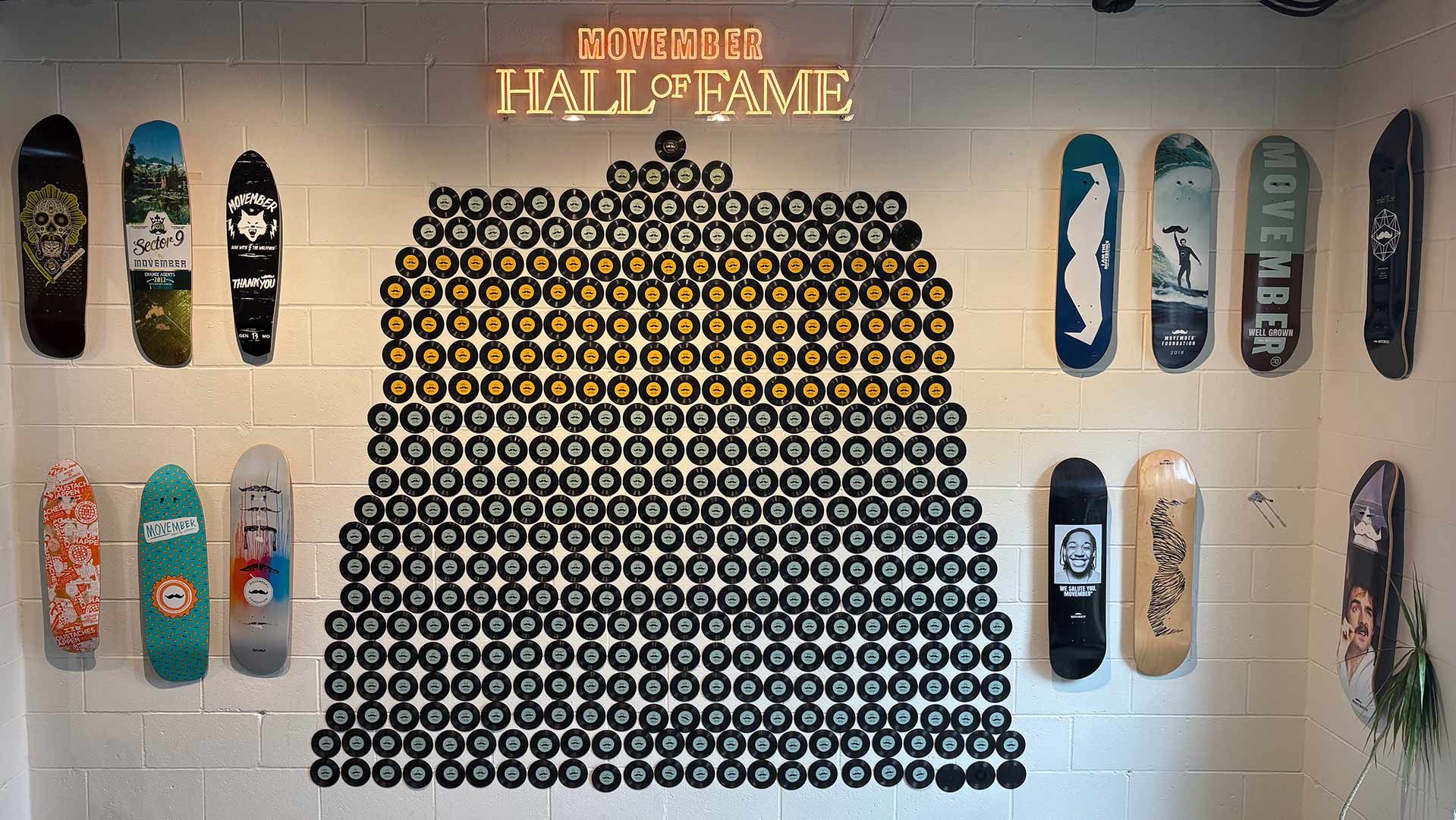 Photo of Movember Hall of Fame merchandise on a wall. It shows replica 7-inch records and Movember-branded skateboards