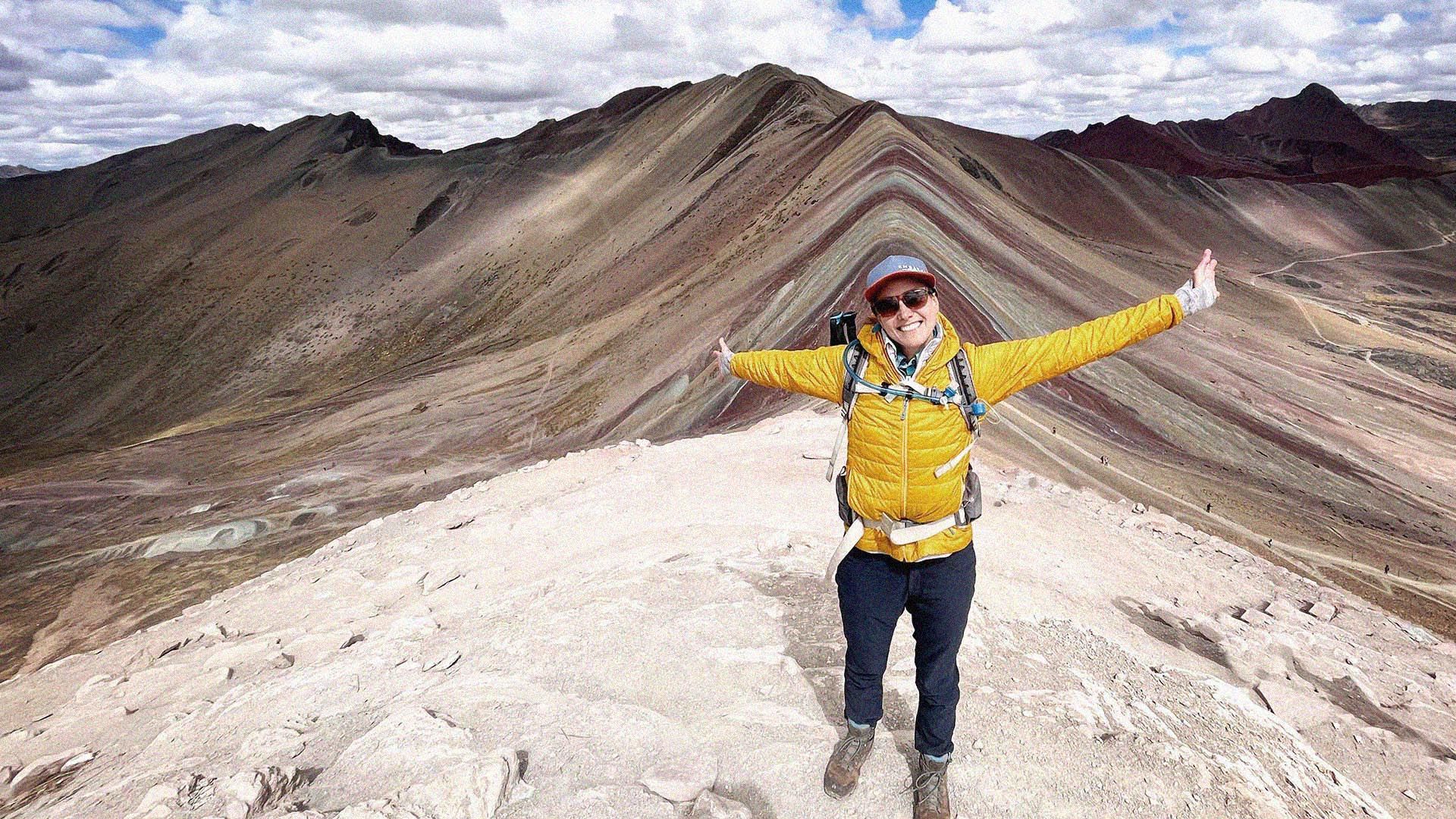 Hiker in cold-weather camping gear posing to camera. The resplendent swirly colours of Peru's Rainbow Mountain are visible in the background.