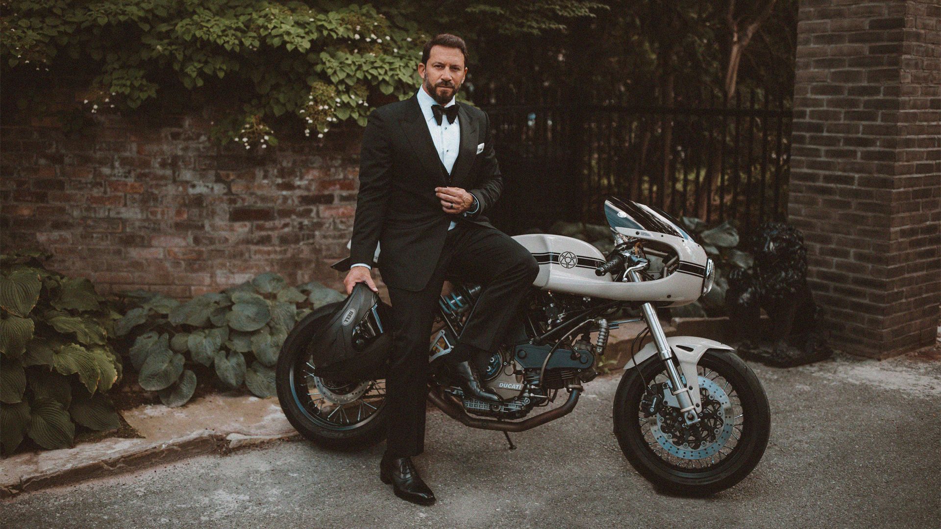 A dapper man dressed in a tux, leans on his parked motorcycle