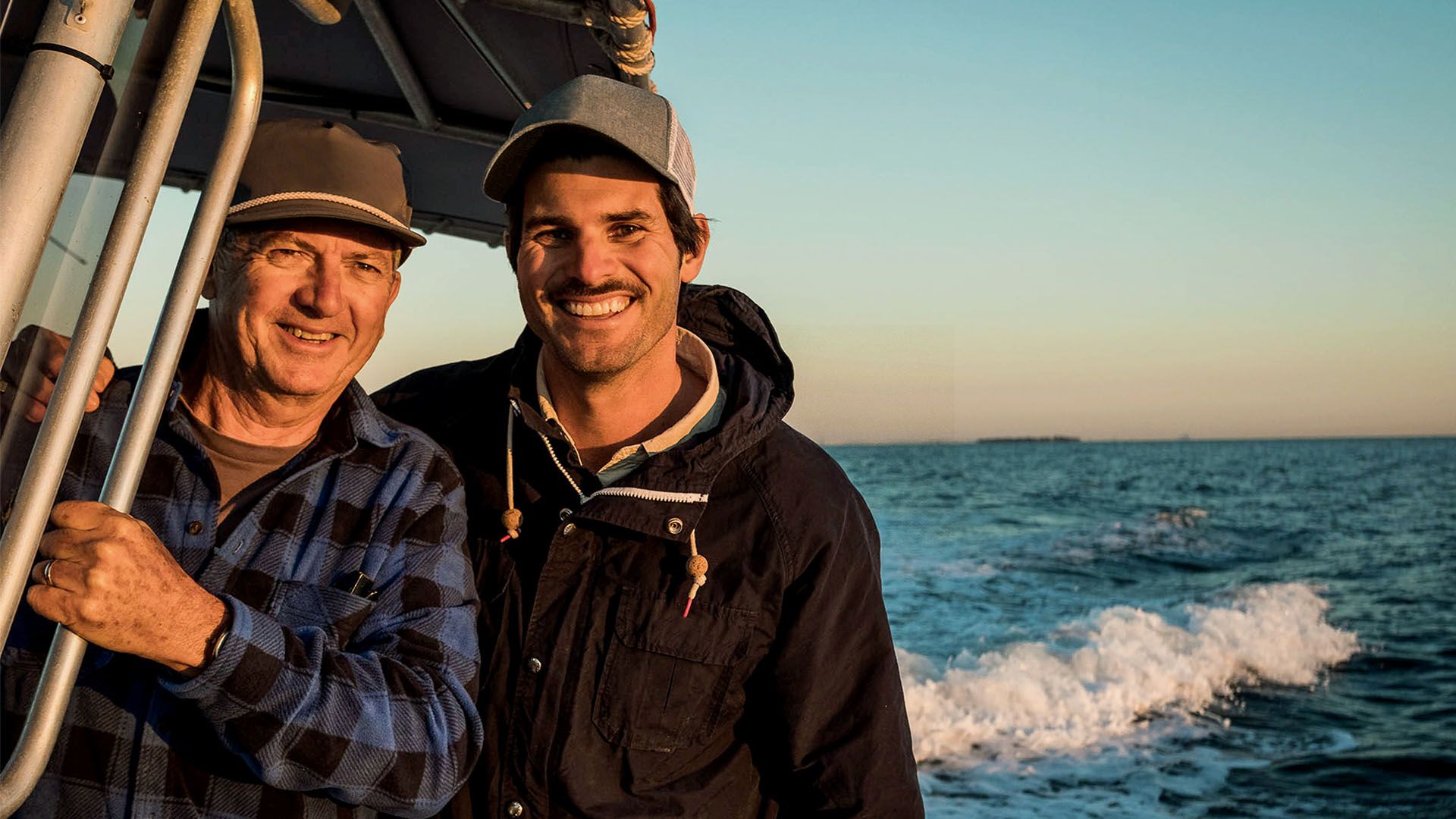Two men stand on fishing boat smiling at camera with ocean in background