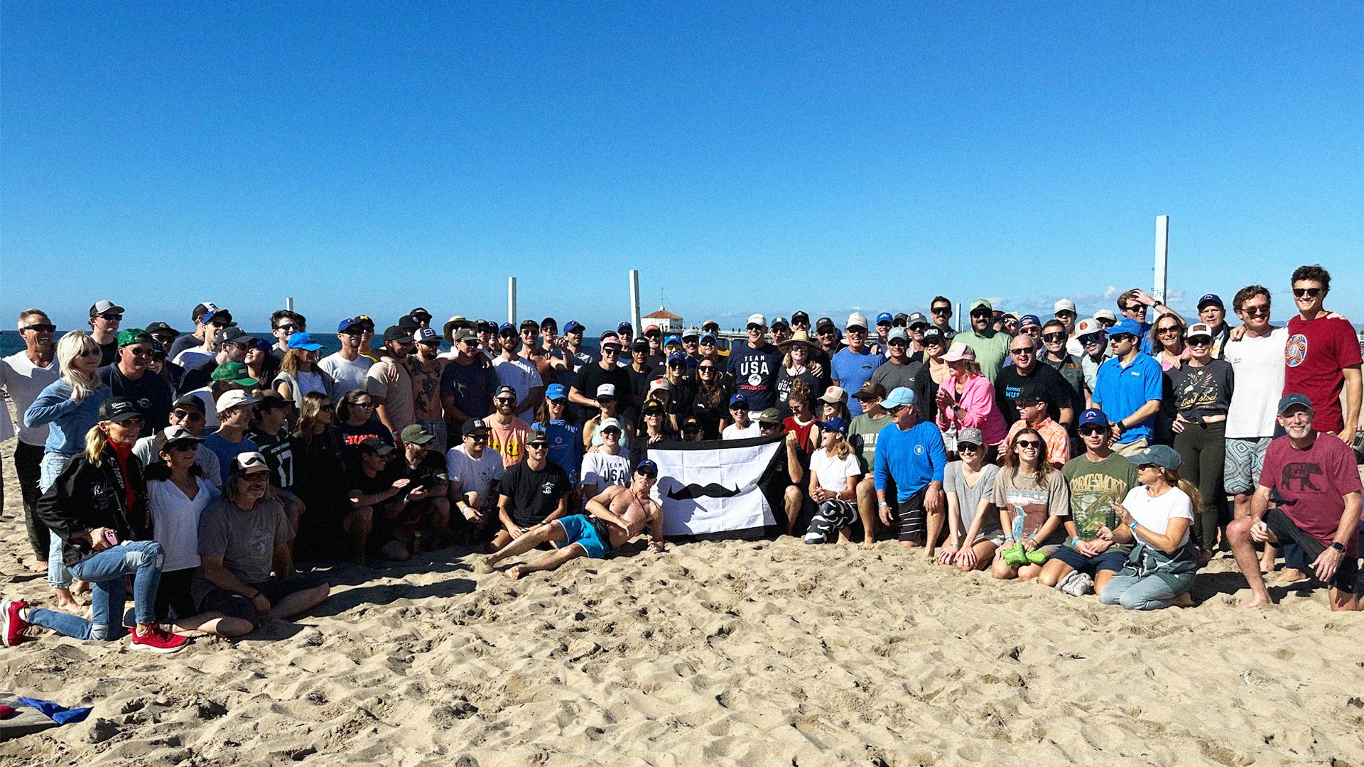 Photo of a crowd posing for a photo at a beach, holding a Movember banner.